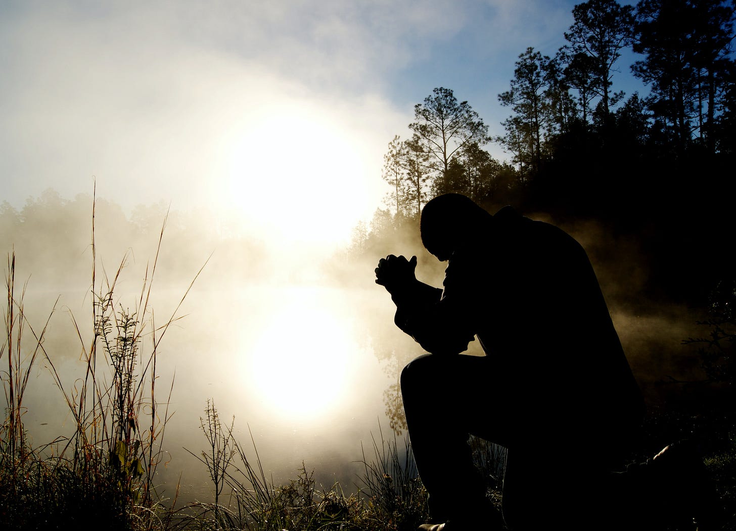 A man is here shrouded in shadows, on one knee in prayer. It's impossible to tell race, and tbh I'm just guessing on gender. He's in a grassy, forested area and is facing toward a pond, over which a bright sun is rising or setting. The sky is blue and clouds are out and shit