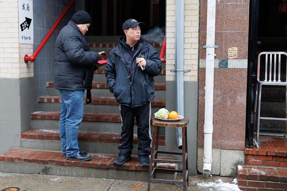 Two men outside a Chinatown building, one smoking a cigar next to a small table with oranges and lettuce for a New Year’s offering. Two men outside a Chinatown building, one smoking a cigar next to a small table with oranges and lettuce for a New Year’s offering.