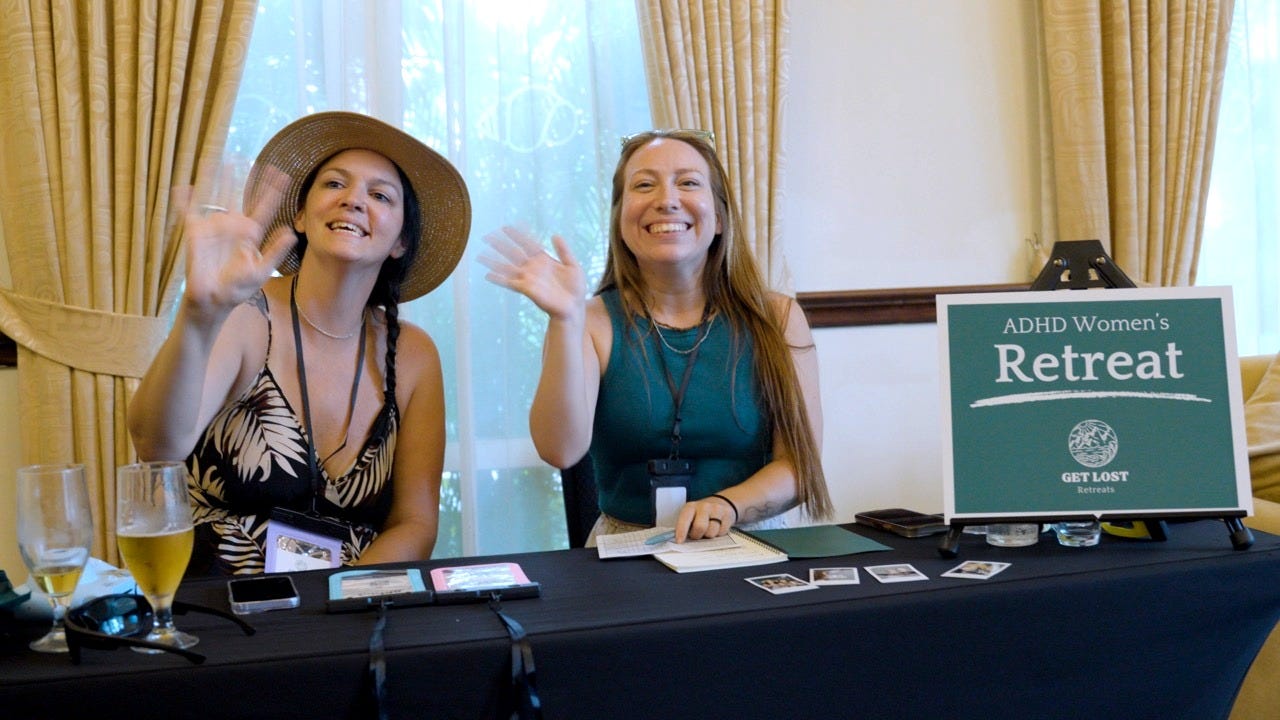 Katie and Trina sit smiling and waving at a welcome table. A sign says "ADHD Women's Retreat" Katie and Trina sit smiling and waving at a welcome table. A sign says "ADHD Women's Retreat"