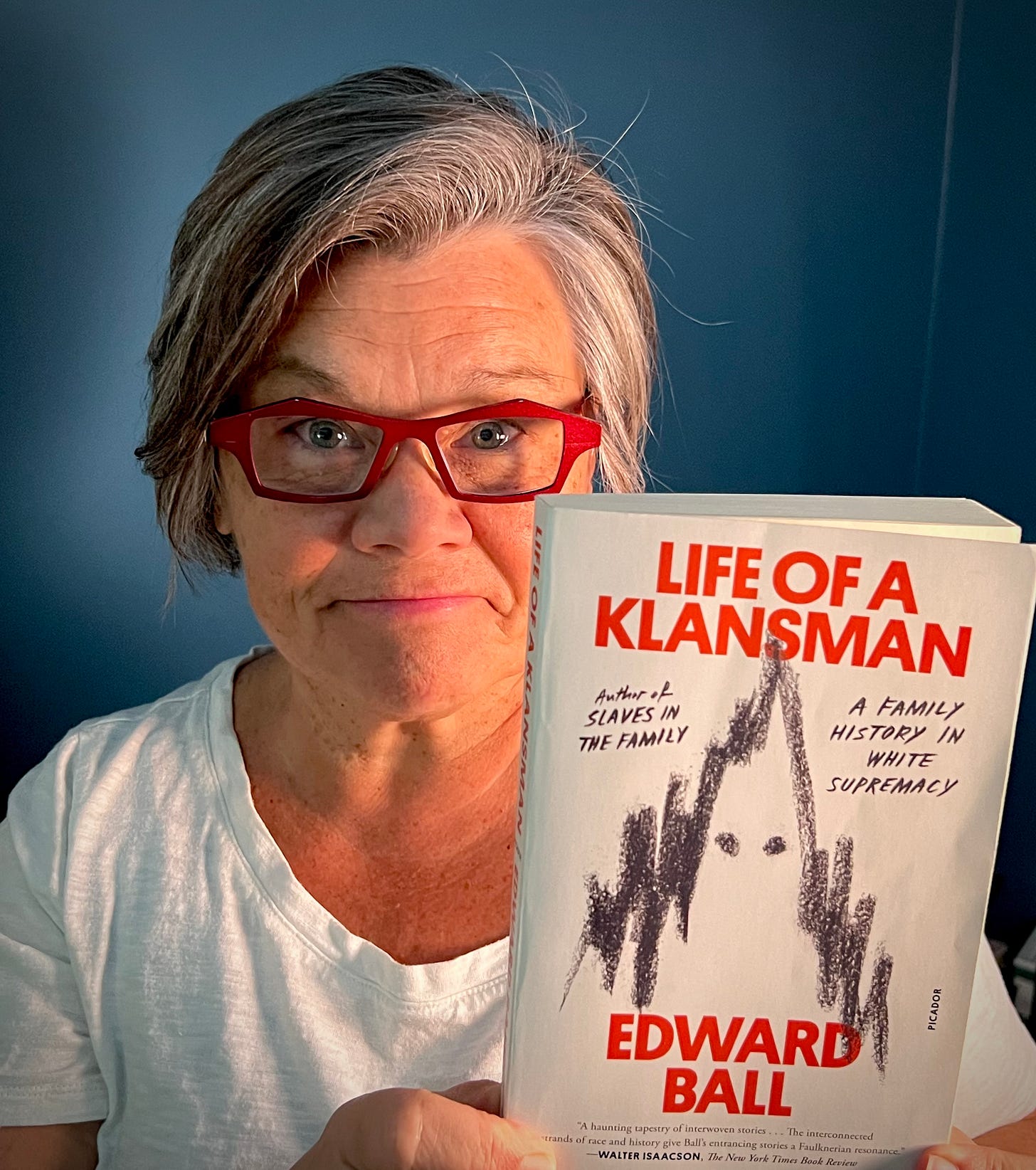 Tamela Rich holding the book, "Life of a Klansman" by Edward Ball. She wears a white tshirt and red glasses, standing before a blue wall