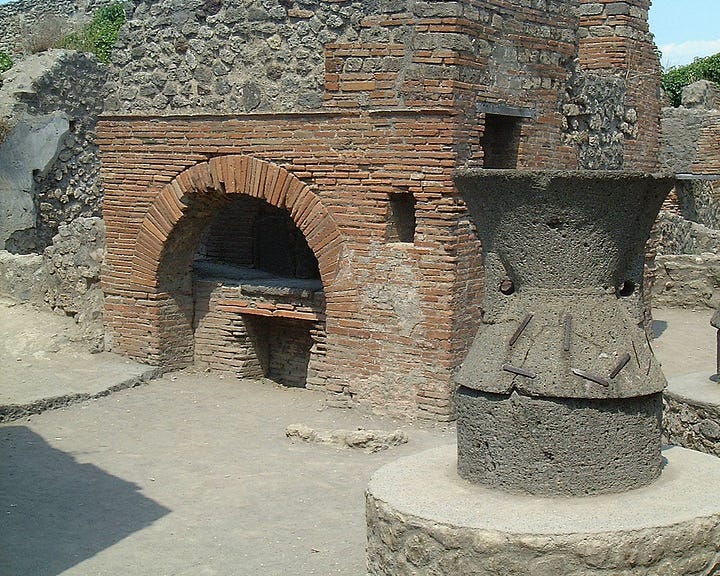 Two Roman grindstones, one form Ostia, one from Pompeii. Nearly the height of the author as the first picture shows with her standing next to a grindstone. Several brick walls stretch away in the left hand picture's background, a fine arched brick oven in the right hand picture.. 