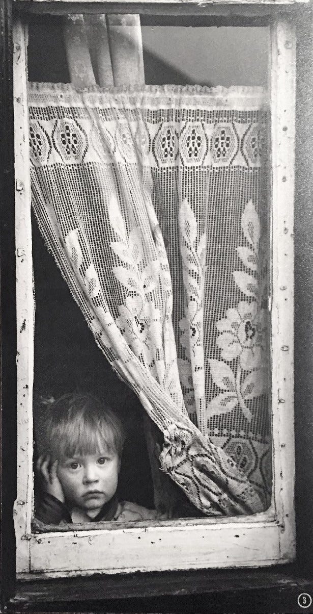 Black and white photo taken in 1962 by Shirley Baker of a similar early twentieth century terrace house bay window with white net curtains in the window. A small White boy is staring towards the camera with the curtain peeled to the side. The photographer has captured the moment beautifully.