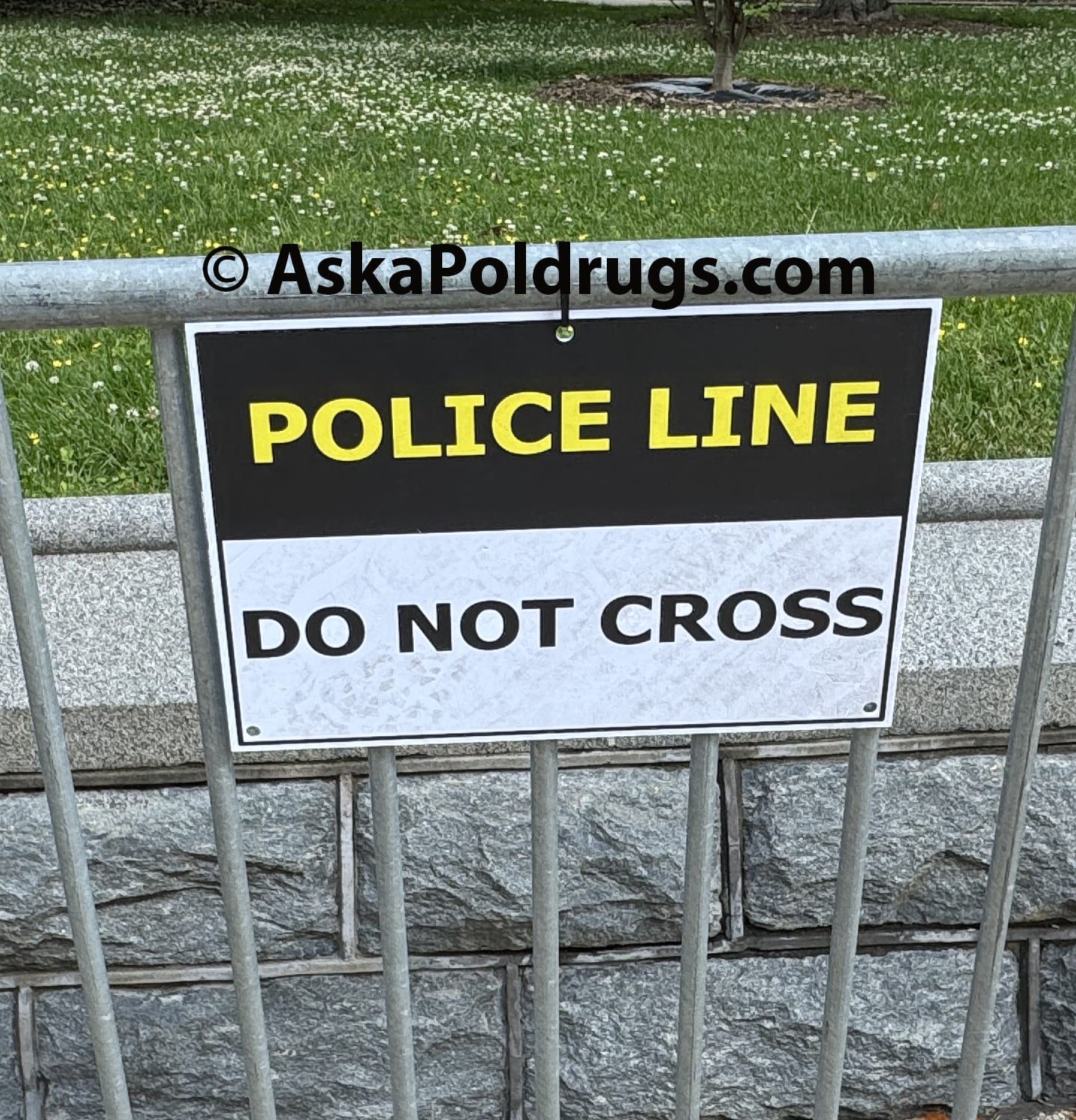 Sign reading ‘Police Line Do Not Cross’ on a metal barricade in U.S. Capitol park with grass and flowers - © AskaPoldrugs.com