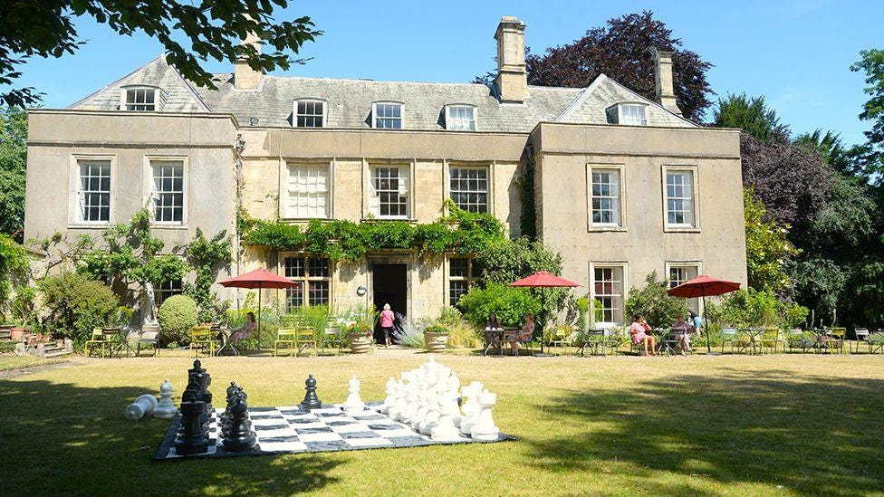 The front of Grantham House a three-storey tan stone building with large windows and vegetation growing around the door and windows. On the lawn in front of the house are people sitting at tables under umbrellas and a giant chess set in the foreground