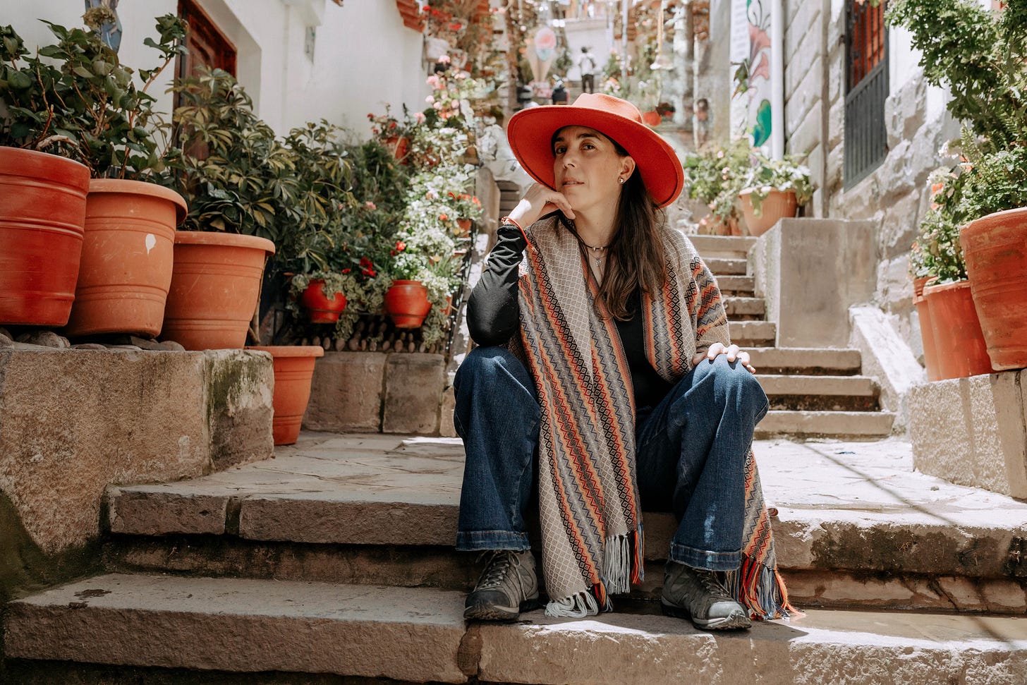 Lorena Saavedra Smith sitting on stone steps in a Peruvian alleyway, wrapped in an Andean shawl and red hat, reflecting on identity and the experience of living between cultures.