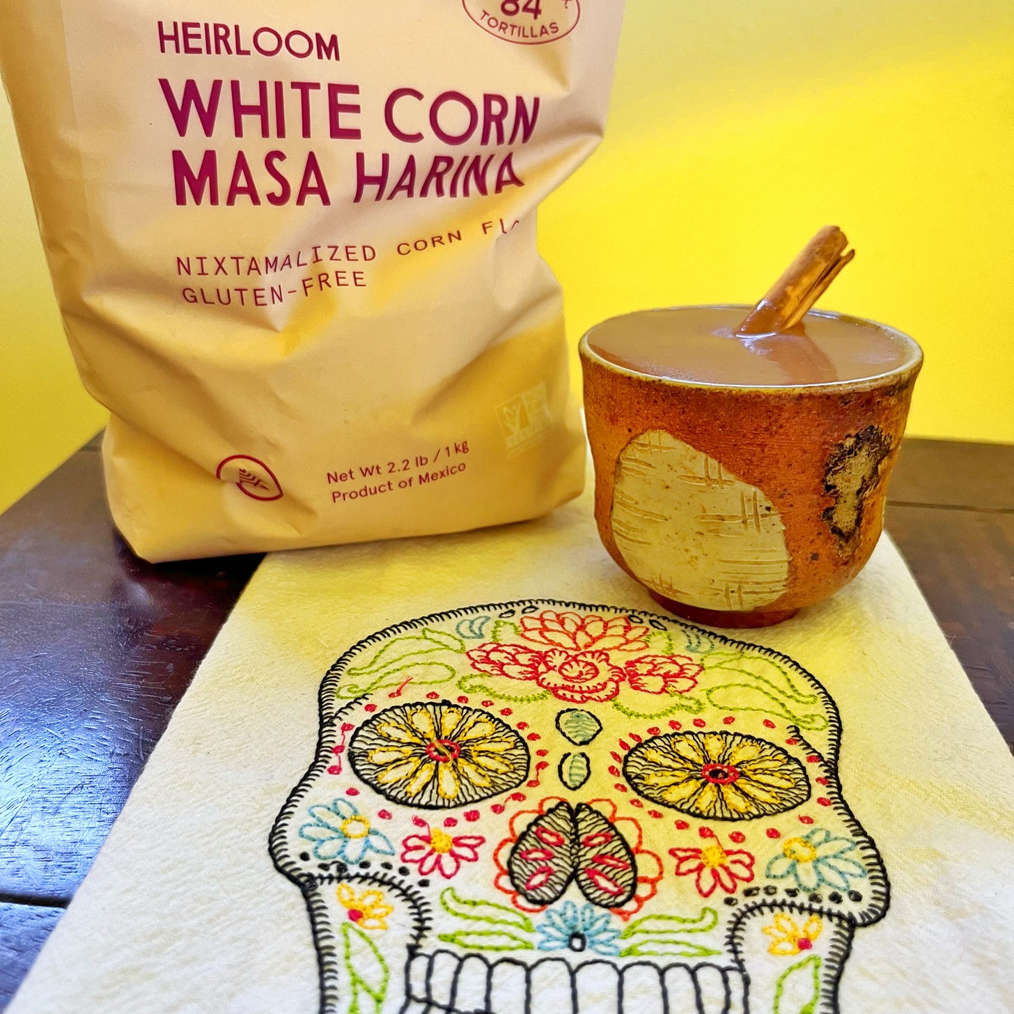 Bag of masa, cup with Champurrado and cinnamon stick, and tea towel with candy skull embroidery
