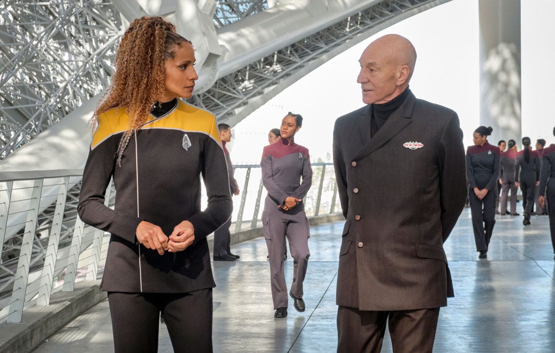 This image shows Commander Raffi Musiker (played by Michelle Hurd, on the left) talking with Admiral Jean-Luc Picard (played by Patrick Stewart, on the right). Several Starfleet cadets are visible walking behind them. This image shows Commander Raffi Musiker (played by Michelle Hurd, on the left) talking with Admiral Jean-Luc Picard (played by Patrick Stewart, on the right). Several Starfleet cadets are visible walking behind them.