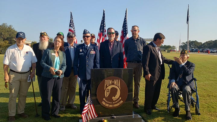 A group of veterans and community members pose outdoors at a POW*MIA memorial. In the foreground is a black granite monument engraved with the POW*MIA emblem and the words “YOU ARE NOT FORGOTTEN,” with a small American flag at its base. Several U.S. flags stand behind the group. Some people wear military dress uniforms and VFW-style caps; one cap reads “Tallahassee,” and another hat bears the U.S. Air Force insignia. On the right, an elderly veteran in a wheelchair salutes while a man in a suit speaks with him. The scene is on a sunny day in a grassy field with cars and flagpoles in the background.