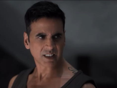 A still from Hindi film, ‘Bhooth Bangla’. Arjun (Akshay Kumar) in a close-up, wearing a dark sleeveless top and looking sharply to one side with a tense, angry expression. The background is dim and out of focus.