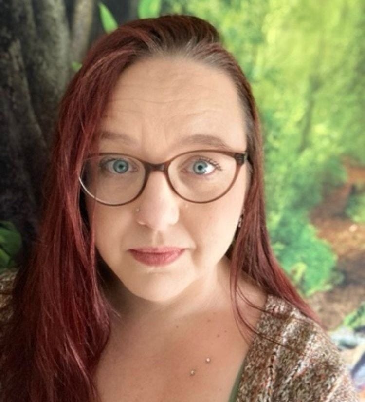 Helen Edgar of Autistic Realms. Helen is standing in front of a woodland background. She has brown glasses, piercings and dyed hair.