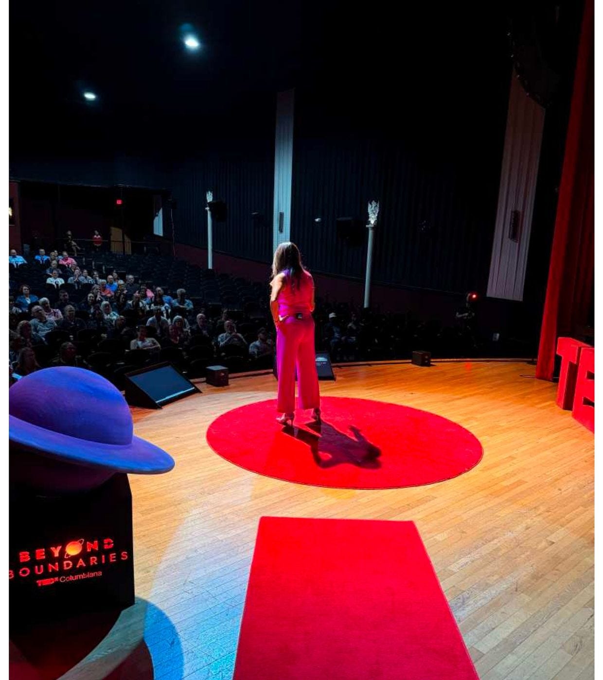 Seen from the wings, a speaker in pink stands on the red circle facing a full house at the Columbiana Arts Theater. A red carpet runner leads to the dot and the big red TED letters stand at the edge of the stage. Seen from the wings, a speaker in pink stands on the red circle facing a full house at the Columbiana Arts Theater. A red carpet runner leads to the dot and the big red TED letters stand at the edge of the stage.