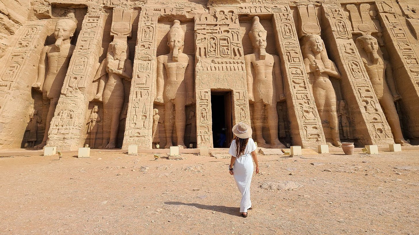 A woman in a white dress and hat walks toward the entrance of an ancient Egyptian temple with large statues carved into the facade.
