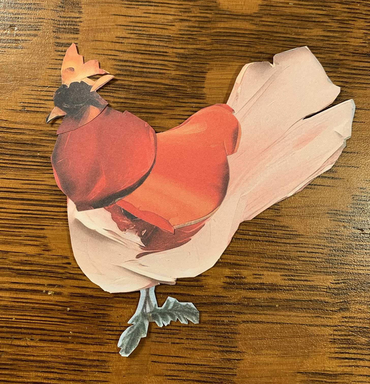 A paper chicken with a pale pink body, orange/red wings, a dark head, and curly green legs. on a wooden background.