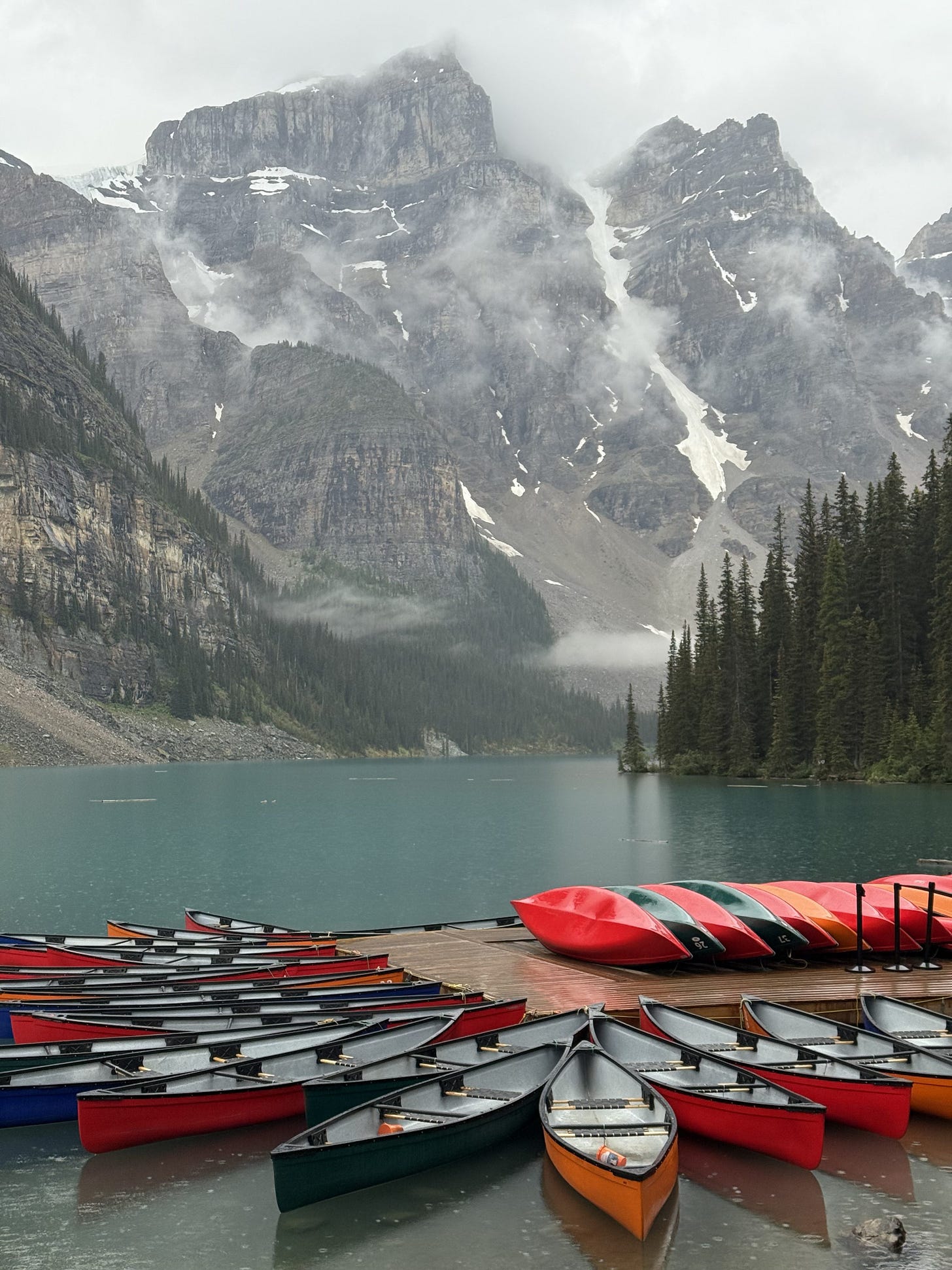A scenic view of colorful canoes lined up at a dock by a misty lake, surrounded by towering mountains and pine trees.