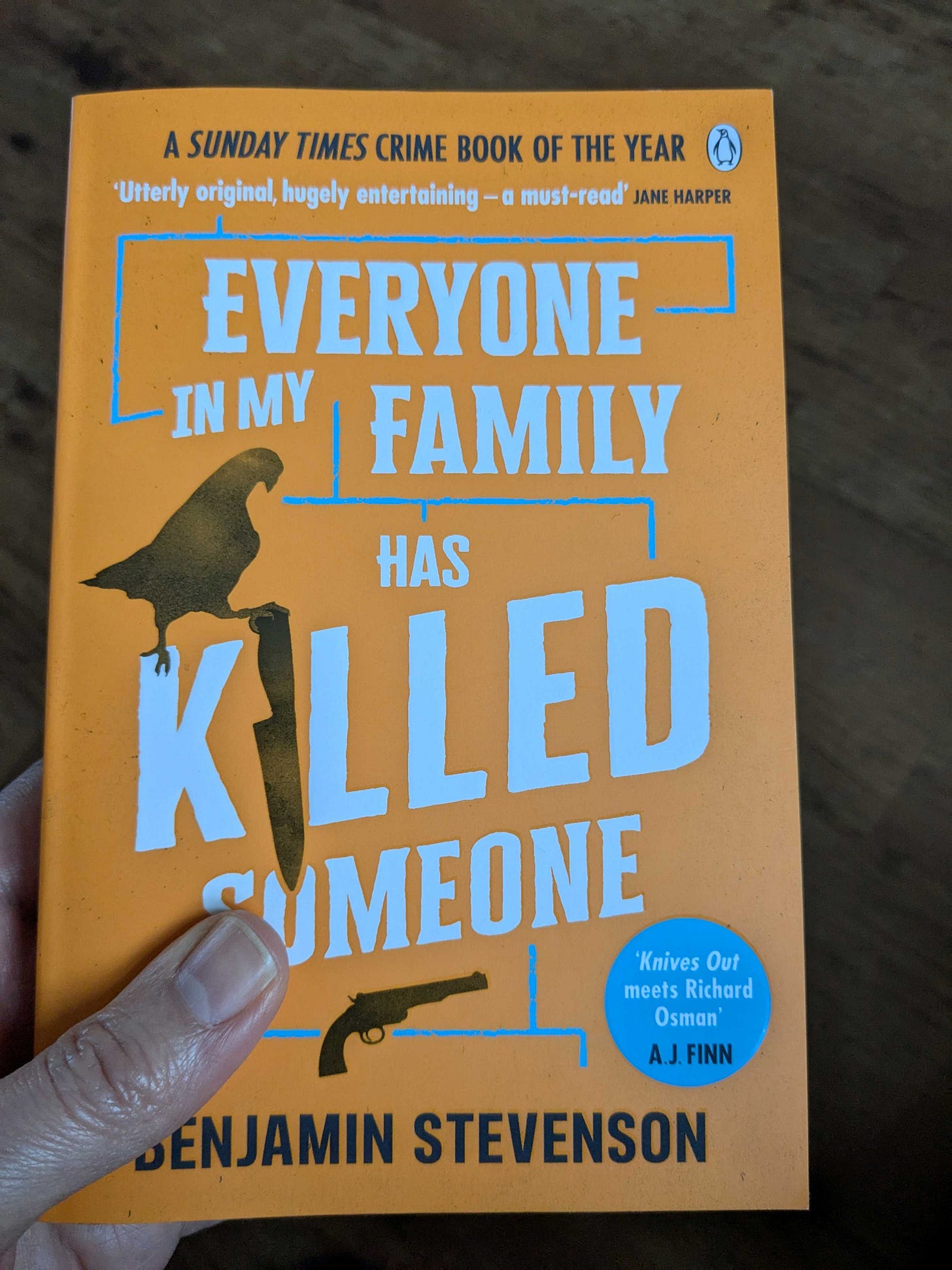 Photo of the book 'Everyone in My Family Has Killed Someone' by Benjamin Stevenson. Cover is orange with a bird, knife and gun on it.
