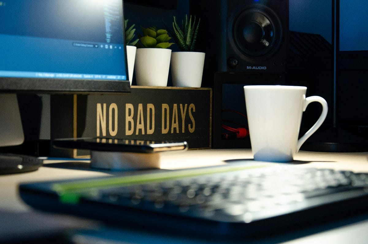 desk in home office with phone, keyboard, white cup, computer screen and speaker. Singing says: “No bad days” — unsplash
