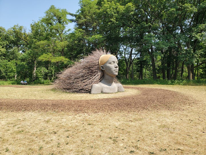 A sculpture of a woman's face and shoulders coming out of the ground, with hair of branches flowing behind her