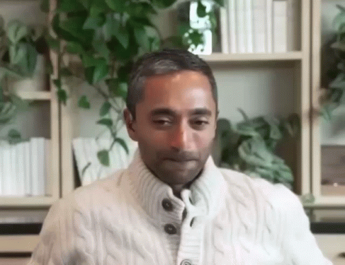 a man in a white sweater is sitting at a table in front of a bookshelf . a man in a white sweater is sitting at a table in front of a bookshelf .