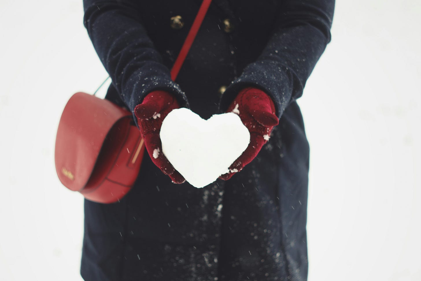 Person holding a heart-shaped snowball. They wear dark red gloves with a matching purse, and a dark coat with brass buttons.