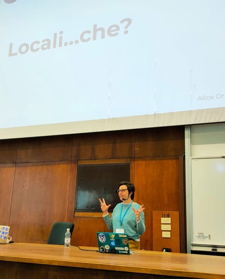 Alice in piedi davanti allo schermo del WordCamp Italia 2022 mentre parla di localizzazione, gesticolando come suo solito. Sullo schermo si legge, in grande, "Locali... che?" Alice in piedi davanti allo schermo del WordCamp Italia 2022 mentre parla di localizzazione, gesticolando come suo solito. Sullo schermo si legge, in grande, "Locali... che?"