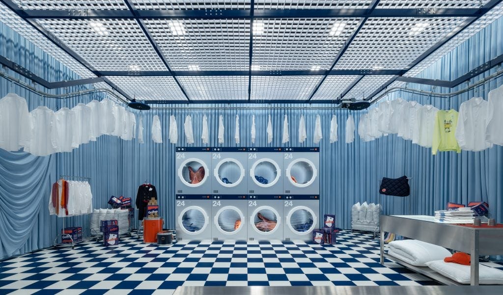 A whimsical laundry room in blue and white tones with checkered floors, curtained walls, and suspended shirts. Oversized dryers are filled with Hermès pieces and detergent boxes labeled “Galop.” Photograph by Sui Sicong. Courtesy of Hermès. A whimsical laundry room in blue and white tones with checkered floors, curtained walls, and suspended shirts. Oversized dryers are filled with Hermès pieces and detergent boxes labeled “Galop.” Photograph by Sui Sicong. Courtesy of Hermès.