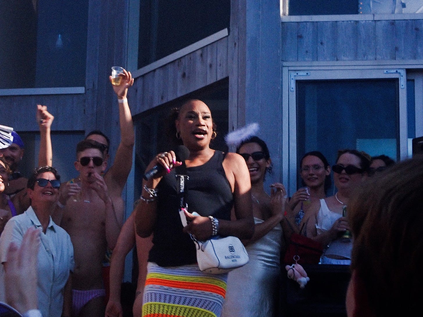 Ceyenne Doroshow, wearing a black top and multicolored skirt, holding a microphone and addressing an excited audience at an outdoor event.