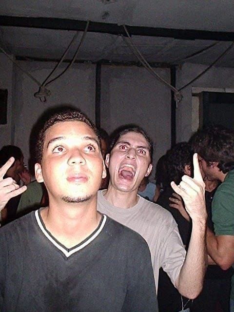 Uma foto antiga em um evento underground, com dois amigos em destaque. À frente, uma pessoa com expressão neutra, olhando para cima, veste uma camiseta preta com gola branca. Atrás, outra pessoa com camiseta branca grita, fazendo gestos de "chifres do rock" com as mãos. Ao fundo, outras pessoas interagem em um ambiente simples, com paredes brancas e cabos pendurados no teto. Uma foto antiga em um evento underground, com dois amigos em destaque. À frente, uma pessoa com expressão neutra, olhando para cima, veste uma camiseta preta com gola branca. Atrás, outra pessoa com camiseta branca grita, fazendo gestos de "chifres do rock" com as mãos. Ao fundo, outras pessoas interagem em um ambiente simples, com paredes brancas e cabos pendurados no teto.