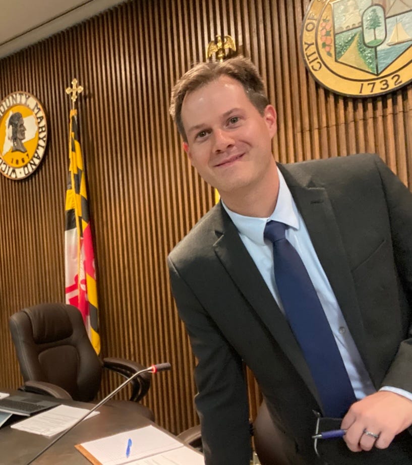 A man in a suit and tie, Josh Hastings, stands confidently in front of a desk.
