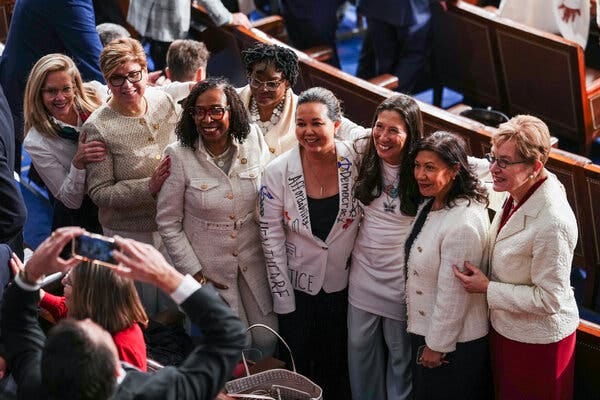 A group of people, many in light-colored clothing, smile while posing closely together. One person wears a white jacket with hand-drawn text. A group of people, many in light-colored clothing, smile while posing closely together. One person wears a white jacket with hand-drawn text.