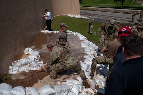 The progression of a sand boil from occurrence to remediation: 1) A soldier in fatigues carries a sandbag downhill alongside a floodway structure (a tall wall) with a cigarette between their lips and eyes turned down toward the target in the grass. The sand boil looks like a gurgling fountain a few inches in diameter, with muddy water spreading around it. 2) The floodway structure, chain of soldiers and ring dyke (several dozen sandbags stacked around the sand boil in a half-moon formation at the base of the wall) are seen from just uphill. A person in a neon yellow safety vest stands in muddy, ankle-deep water as it rises inside the ring dyke, which is now a few sandbags high. Inside the floodway structure, floodwaters as sludgy as brownie mix appear to loom several feet over the heads of the soldiers working on the landside. Two military SUVs pass on the road in the background. 3) Soldiers in fatigues pass sandbags down from pallets on a flatbed. In the foreground, a blond soldier with a square jaw and solemn expression looks on in a camouflage cap. 4) Soldiers in fatigues stand perpendicular to the floodway structure, passing sandbags toward the ring dyke. 5) Two people stand side by side, facing the ring dyke with their backs to the photographer. Muddy water spills over the sides of the ring dyke, which now stands five or six sandbags high. The person on the right is in fatigues. The person on the left wears a neon yellow safety vest with the red and white turreted castle emblem of the U.S. Army Corps of Engineers. 6) Several members of the Oklahoma National Guard stand in, on and around the ring dyke in fatigues. One is submerged to calf- or knee-height in the muddy water spilling over the edges of the ring dyke. Another steps calf-deep into the pool with one leg, straddling the ring dyke to position a sandbag.