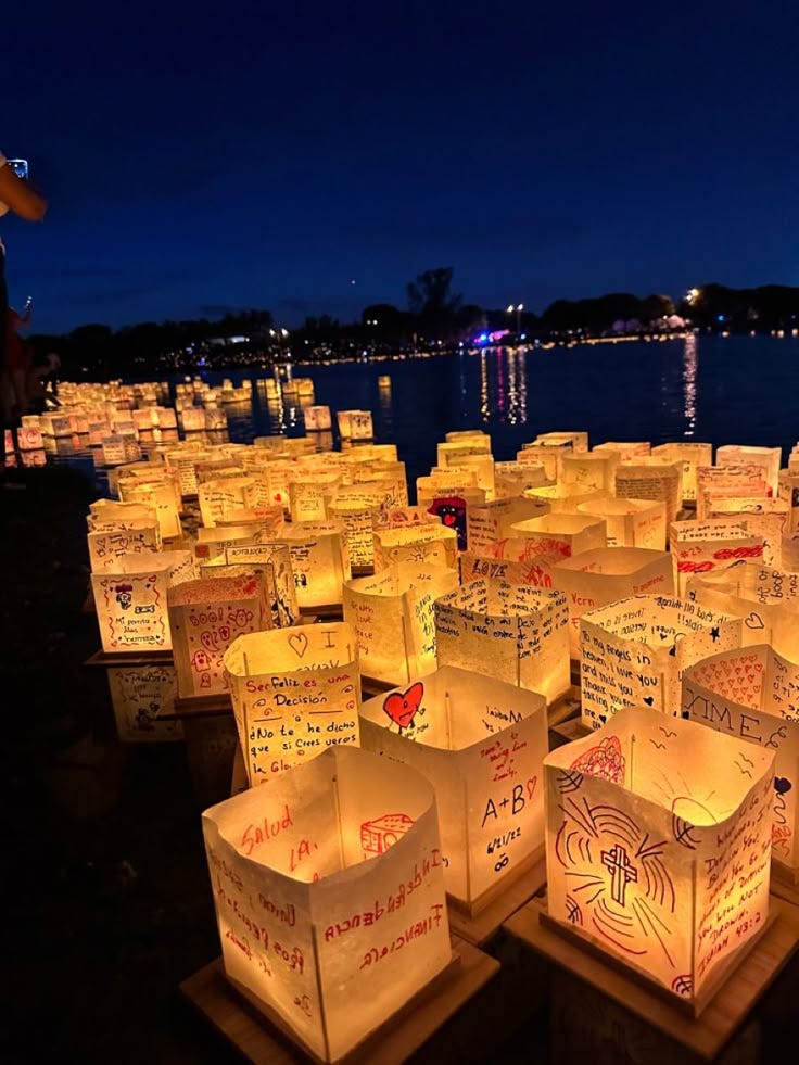 This may contain: many lit up boxes are sitting on the ground near water at night with people standing around them