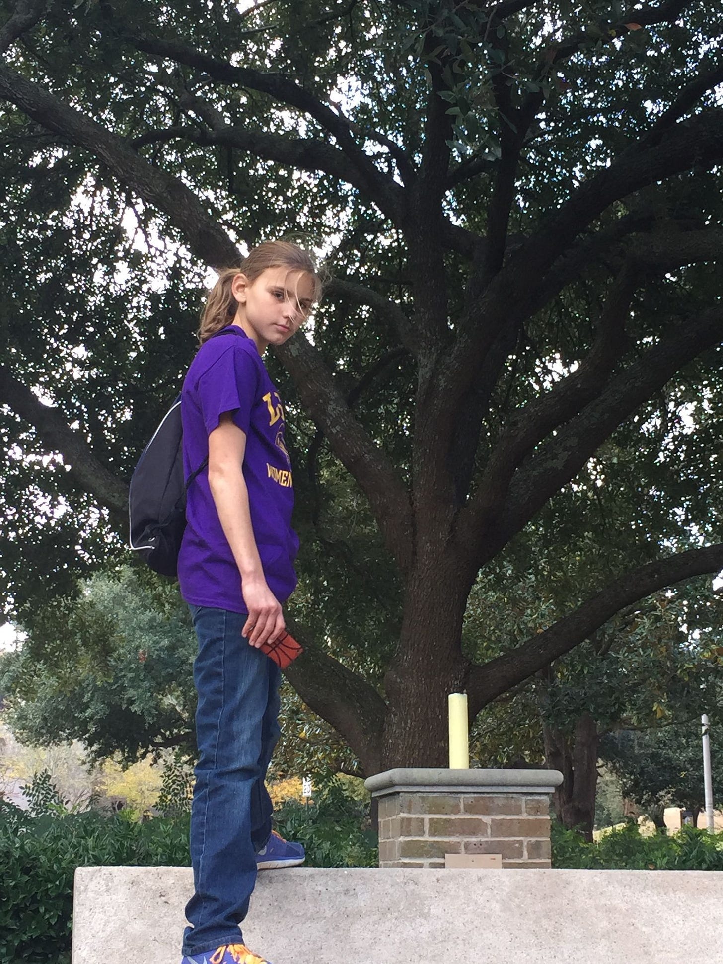 Myself at age 11, standing in front of a tree and looking sideways at the camera. I am wearing baggy jeans, a purple t-shirt, and my dirty-blonde hair is up in a tight ponytail.