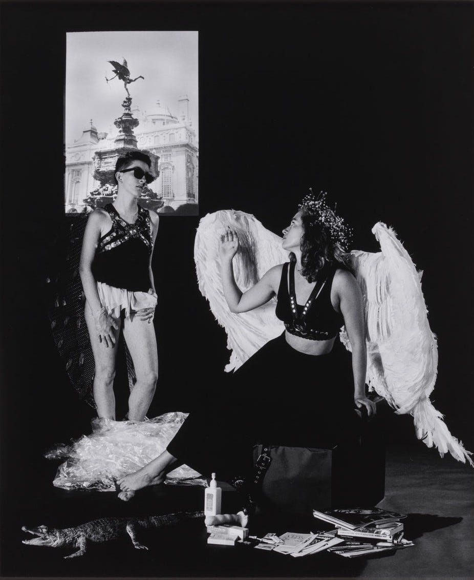Two people in elaborate costumes face each other. One is sitting wearing large, white angel wings, while the other stands. A statue photo is visible behind the person who just emerged from the clingwrap. The clingwrap sits around their feet on the ground, next to the faux alligator. The person who was wrapped up is wearing a leather chest harness, a long frilly shirt covering their crotch, sunglasses, and latex gloves on both hands. They also have dark angel wings, with pieces of metal hanging off of them in several places. The person sitting down is looking over their wings to look at the other angel. The floor in front of them is still full of magazines, pamphlets, books, and a dildo with some lotion/lube. This photo is in black and white.