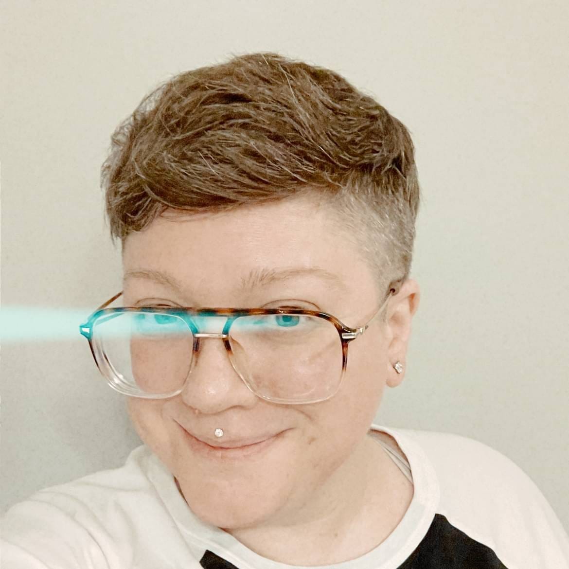 Picture of a white person smiling, with short, brown and grey hair, large-framed tortoiseshell glasses like their dad probably wore in the 1980s, and a medusa piercing. The photo has been edited to wash the picture out and the subject has a beam of pale blue light cutting across their eyes like a laser. This person's kids definitely think they are very cool and up on current trends. This person's cats respect them completely. Picture of a white person smiling, with short, brown and grey hair, large-framed tortoiseshell glasses like their dad probably wore in the 1980s, and a medusa piercing. The photo has been edited to wash the picture out and the subject has a beam of pale blue light cutting across their eyes like a laser. This person's kids definitely think they are very cool and up on current trends. This person's cats respect them completely.