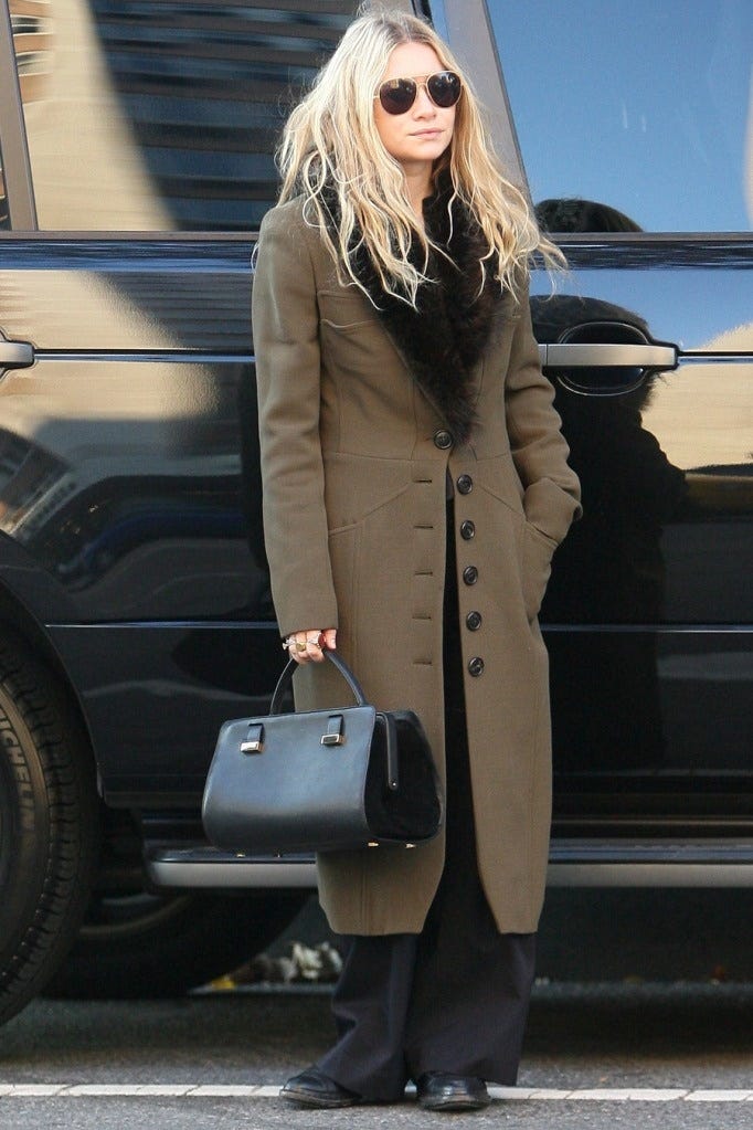 This may contain: a woman with blonde hair and sunglasses holding a black handbag while standing in front of a car