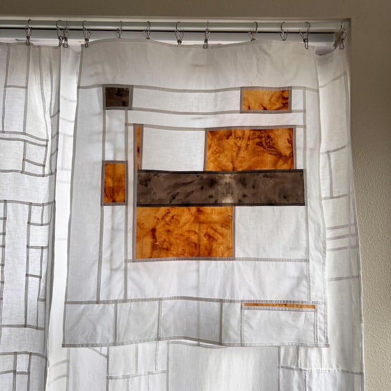 May include: White fabric shower curtain with a geometric design. The curtain features a patchwork of orange and brown fabric squares and rectangles, outlined with gray stitching. The curtain is hanging from a metal rod with clips.