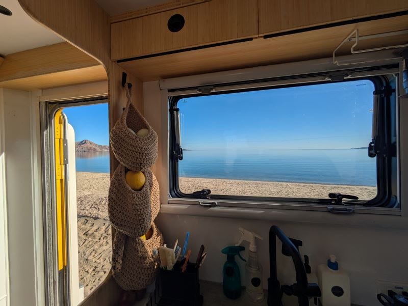 a view of a calm blue sea beneath a matching blue sky, with some small mountainous rock formations in the distance, all viewed from inside Walter's Snuggery's kitchen, looking out the window and door