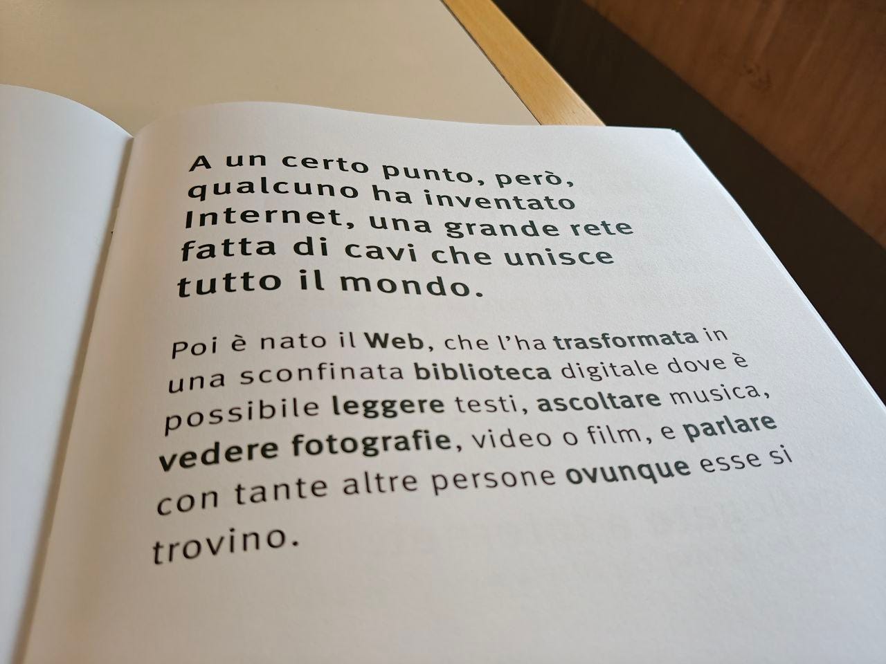 Pagina di un libro aperto con testo stampato in caratteri neri grandi e leggibili. Il testo recita: 'A un certo punto, però, qualcuno ha inventato Internet, una grande rete fatta di cavi che unisce tutto il mondo. Poi è nato il Web, che l'ha trasformata in una sconfinata biblioteca digitale dove è possibile leggere testi, ascoltare musica, vedere fotografie, video o film, e parlare con tante altre persone ovunque esse si trovino.