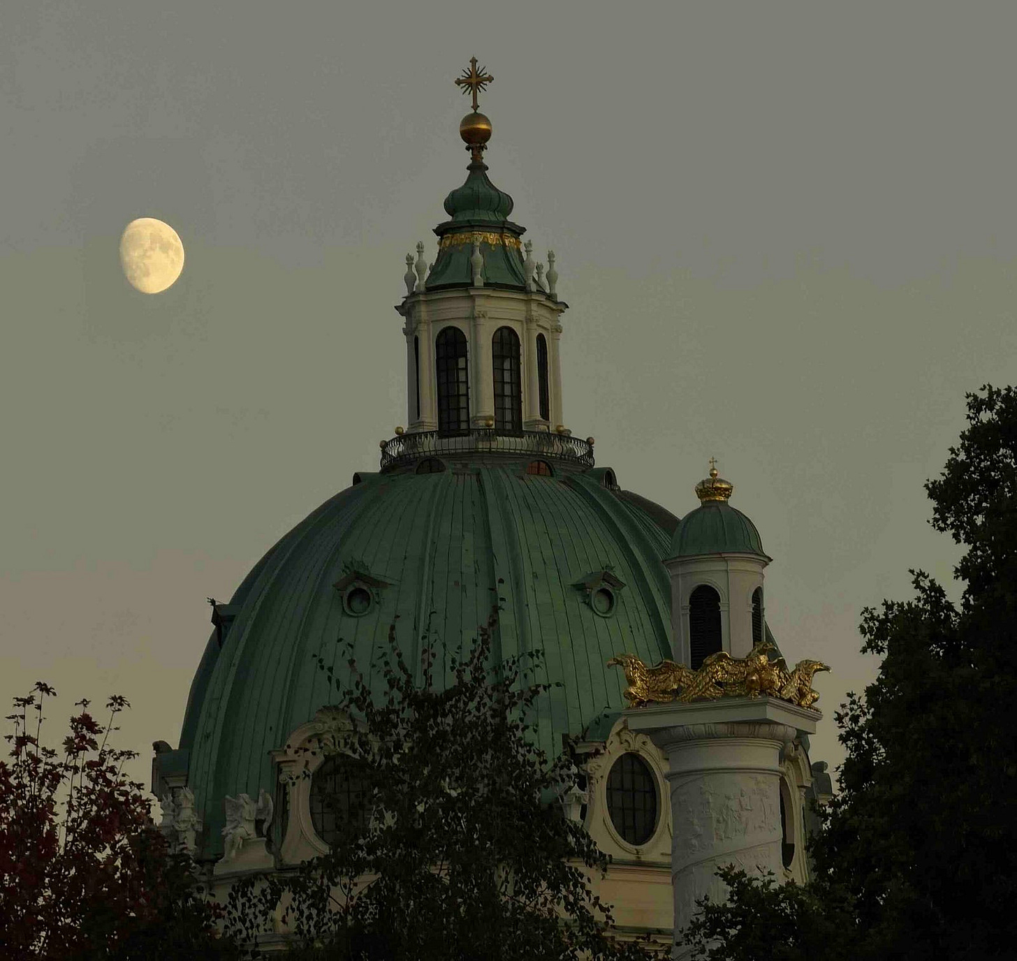 A grand domed building with classical architecture, featuring a green copper dome topped by a cupola and cross, stands partially obscured by trees. Ornate golden embellishments and statues accent the structure, especially on a turret to the right. A nearly full moon glows in the muted gray sky, suggesting dusk or dawn, adding a serene contrast to the building’s opulence. A grand domed building with classical architecture, featuring a green copper dome topped by a cupola and cross, stands partially obscured by trees. Ornate golden embellishments and statues accent the structure, especially on a turret to the right. A nearly full moon glows in the muted gray sky, suggesting dusk or dawn, adding a serene contrast to the building’s opulence.