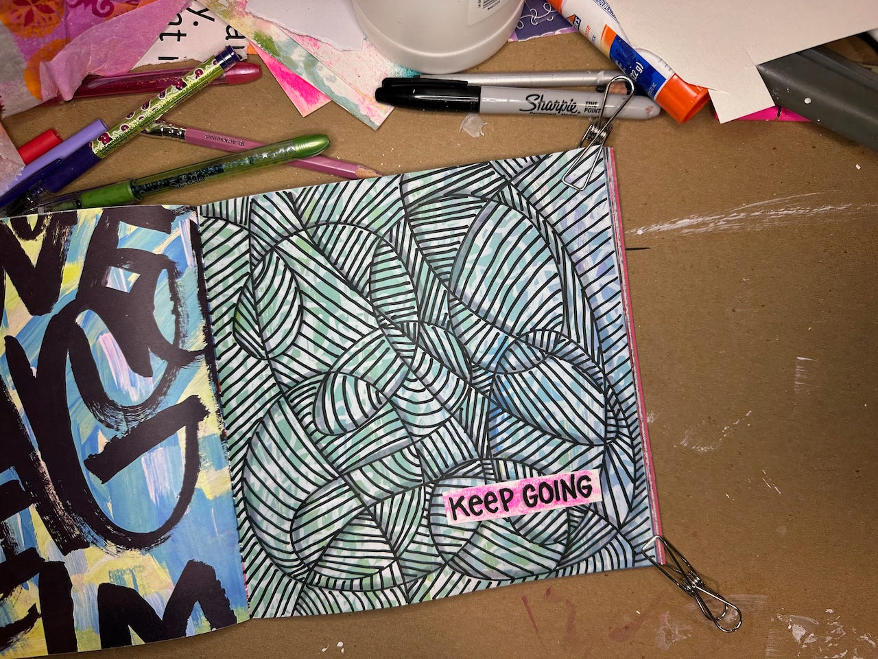 Picture of a messy desktop with an open art journal. The page is covered with a doodle in black lines and the words KEEP GOING on a pink piece of paper on top.