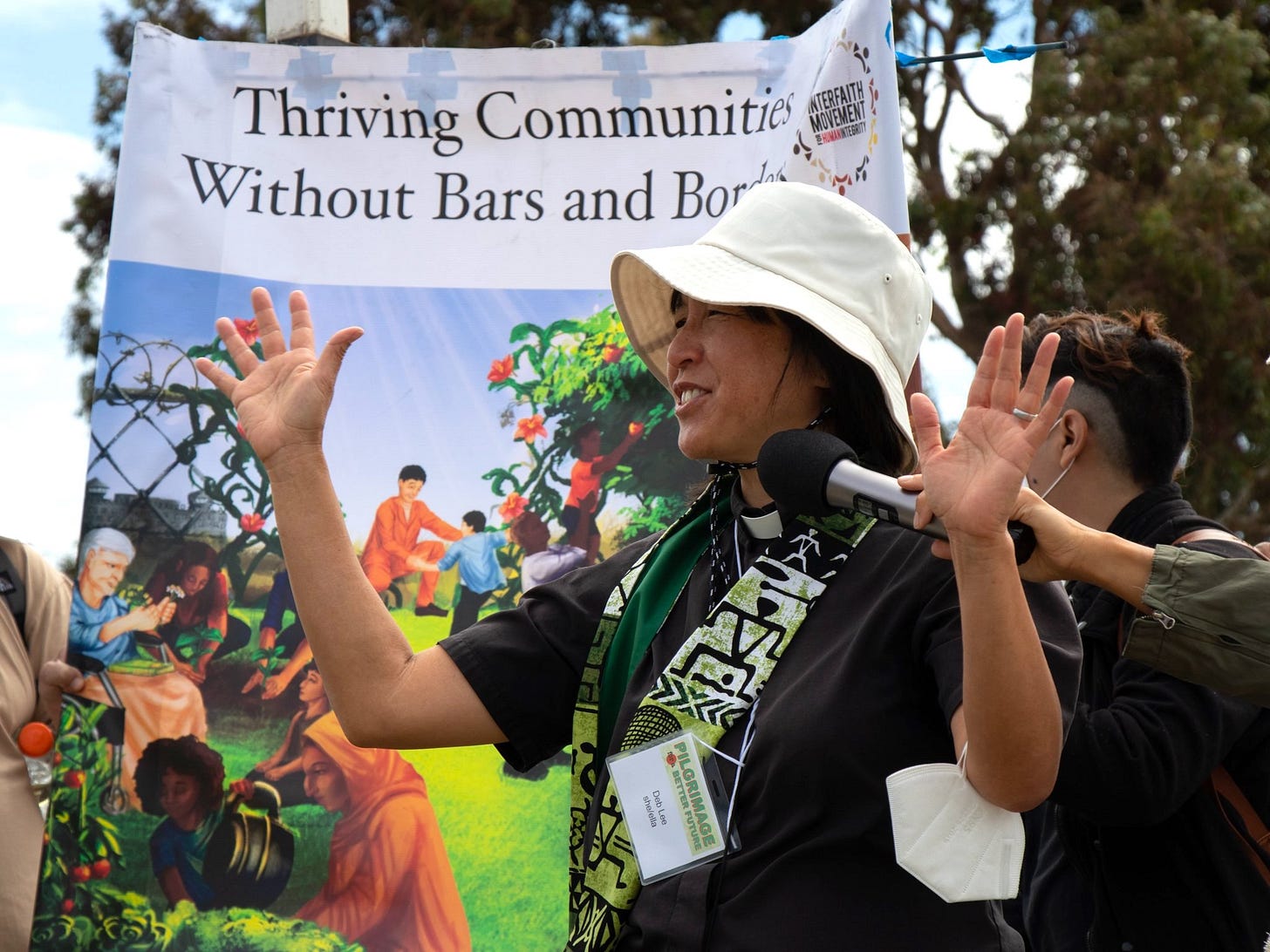 Rev Deb speaks at a rally on a bright sunny day. Behind her is a large banner that reads "Thriving Communities Without Bars and Borders."