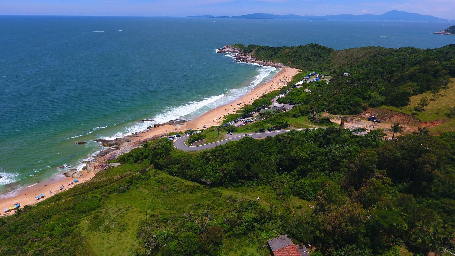 Aerial view of Praia do Pinho showing a long crescent of sandy beach bordered by green hills, a narrow access road with parked cars, small beach structures, and the Atlantic Ocean stretching toward the horizon.