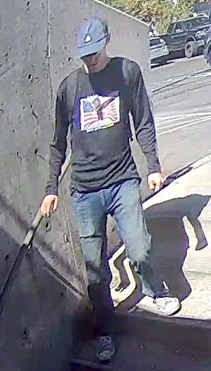 Person wearing a dark long-sleeve shirt with a colorful graphic, blue jeans, and sneakers walking down a sunlit concrete ramp beside a wall.