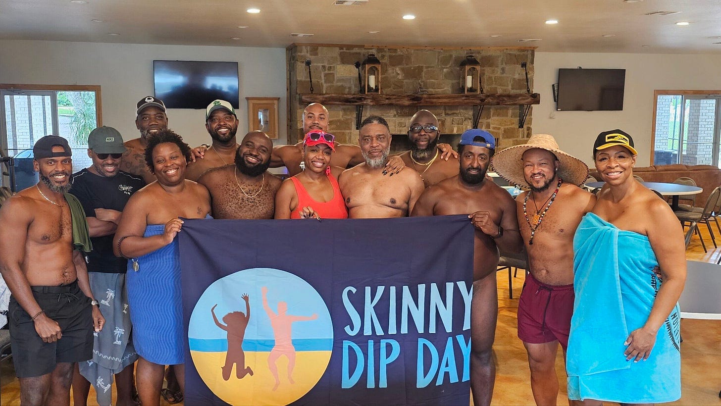 Members of the Black Naturists Association pose together indoors holding a Skinny Dip Day flag, some wrapped in towels and others partially nude, smiling in front of a stone fireplace. Members of the Black Naturists Association pose together indoors holding a Skinny Dip Day flag, some wrapped in towels and others partially nude, smiling in front of a stone fireplace.