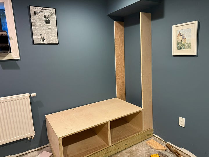 Alt text: A four-photo collage documenting the construction of a custom built-in reading nook. The top-left image shows an empty corner of a room with blue walls, white trim, and a ceiling bulkhead. The top-right image shows the initial framework for a bench seat and a vertical cabinet side installed beneath the bulkhead. The bottom-left image shows the bench structure further along, with additional framing and support panels in place. The bottom-right image shows the nearly completed project, including a built-in bench with storage compartments below and a floor-to-ceiling bookshelf fitted beneath the bulkhead. Together, the images illustrate the progression from an empty corner to a custom built-in seating and storage feature.