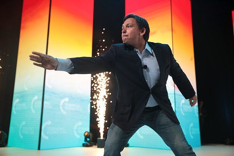 Ron DeSantis, his lips pursed and right arm arm outstretched dramatically, leaning forward a bit as if casting a spell. Behind him, a small onstage prop spits a thin fountain of sparks Ron DeSantis, his lips pursed and right arm arm outstretched dramatically, leaning forward a bit as if casting a spell. Behind him, a small onstage prop spits a thin fountain of sparks