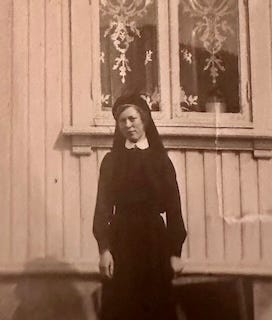 A young woman, dressed in a dark nursing uniform which makes her look a bit like a nun, with a head covering and full dress, with a white collar, cocks her head and shyly smiles at the camera, in front of the side of a white frame house, a window with lace curtains over her shoulder. A young woman, dressed in a dark nursing uniform which makes her look a bit like a nun, with a head covering and full dress, with a white collar, cocks her head and shyly smiles at the camera, in front of the side of a white frame house, a window with lace curtains over her shoulder.