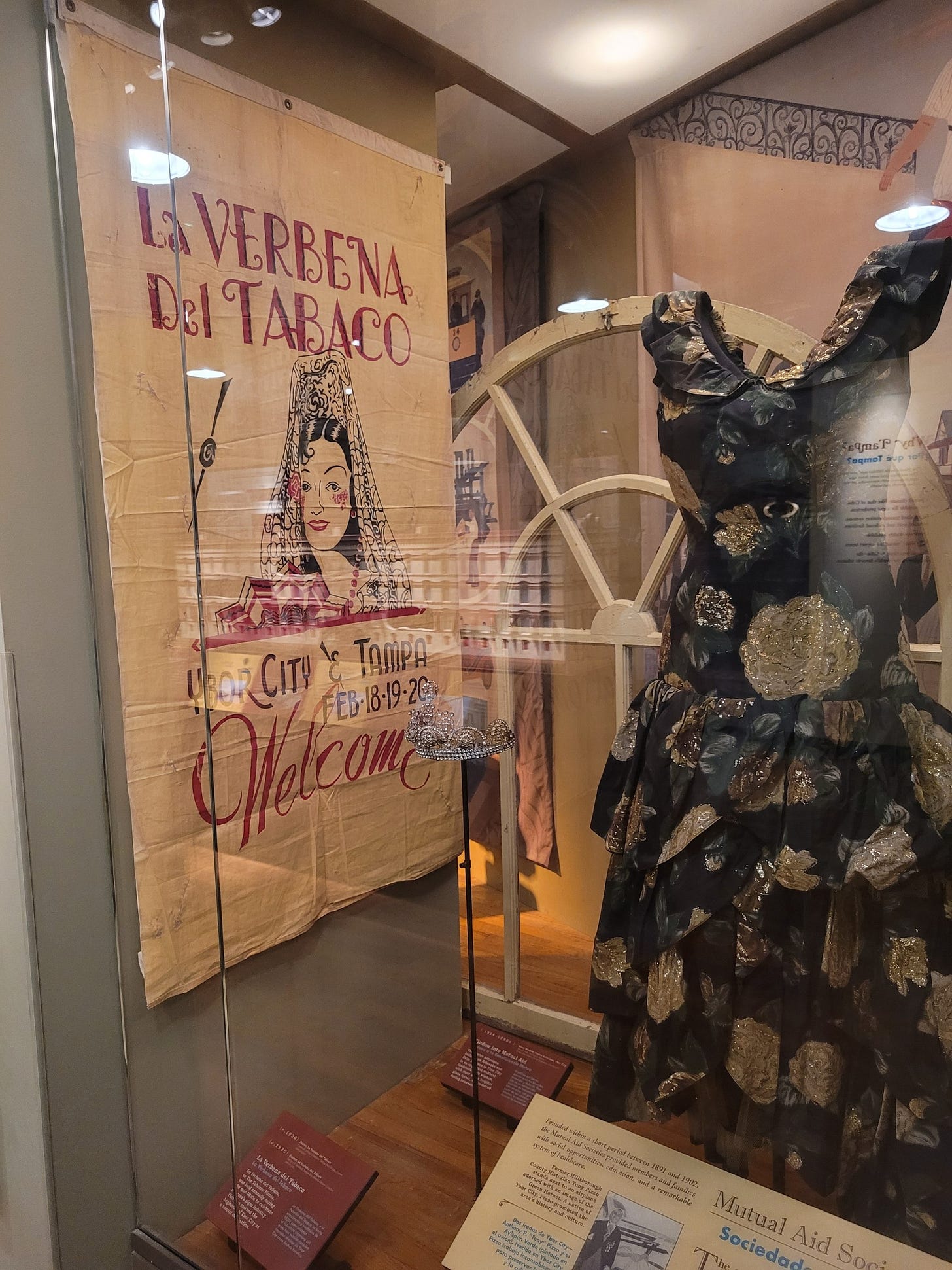 Vitrine de um museu com um cartaz do lado esquerdo escrito La Verbena del Tabaco e o desenho de uma mulher. Na frente, um vestido escuro estampado com alças grossas e sem mangas, talvez na altura dos joelhos.