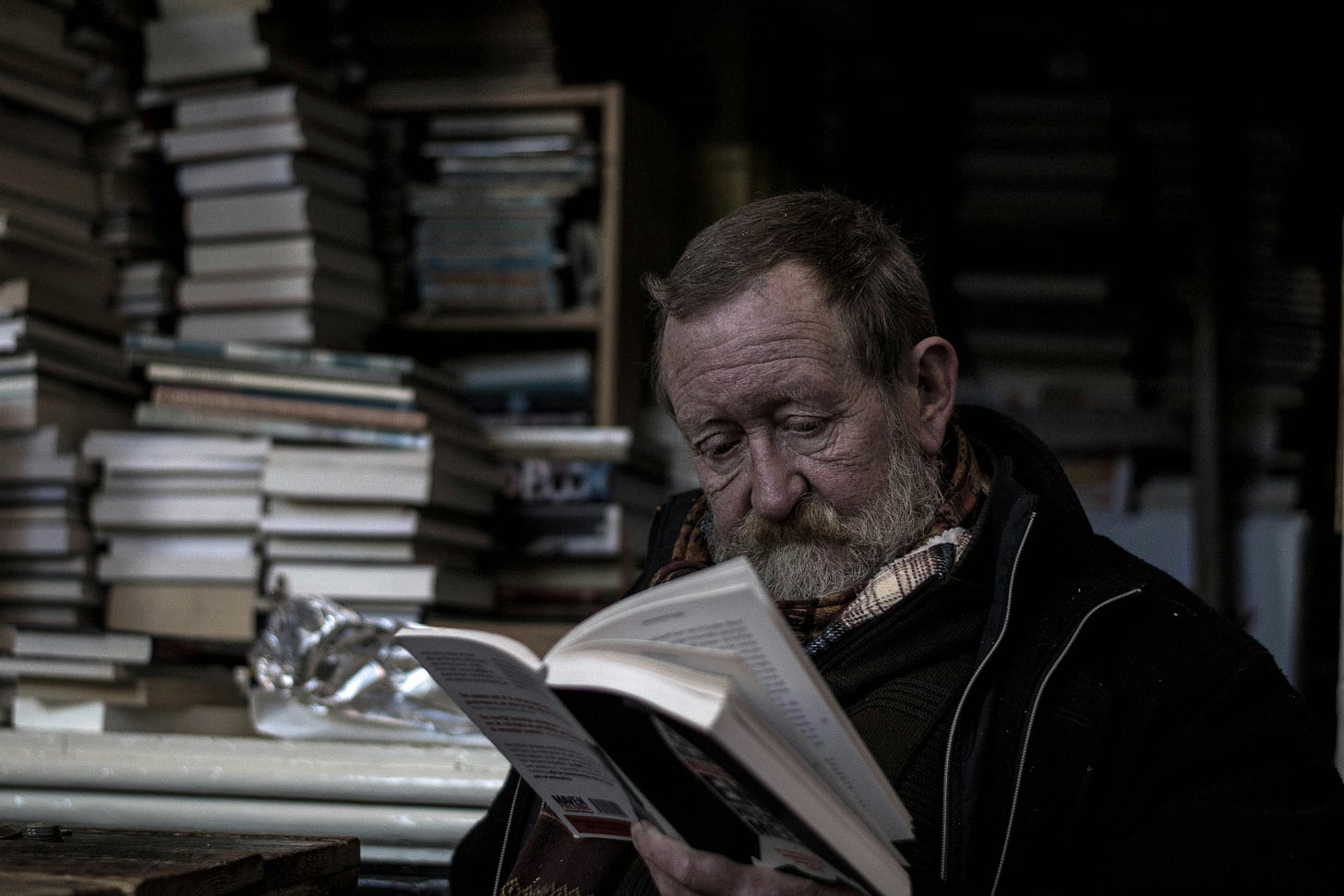 An older man reading a book, with stacks of books on a table in the background