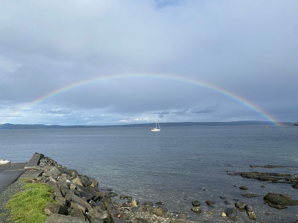 A sailboat sits directly under a full and vibrant rainbow.
